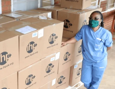 International Health Partners worker in scrubs beside stacked boxes of supplies